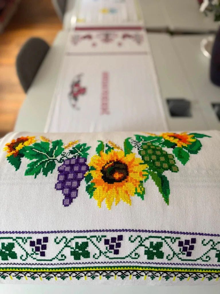 A cross-stitch runner with sunflowers and bunches of grapes on white linen.