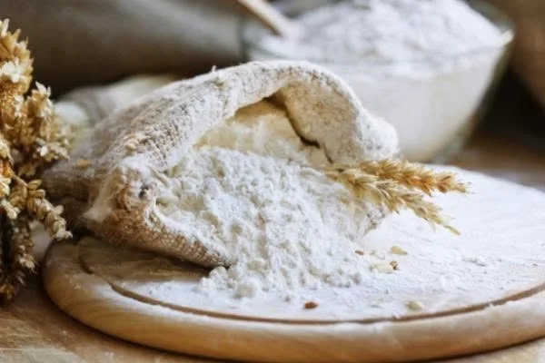 An open sack of flour on a wooden board, with dried wheat beside it.