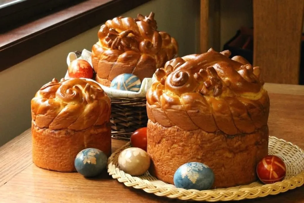 Two tall Easter breads with icing, on a wooden table with painted eggs around them.