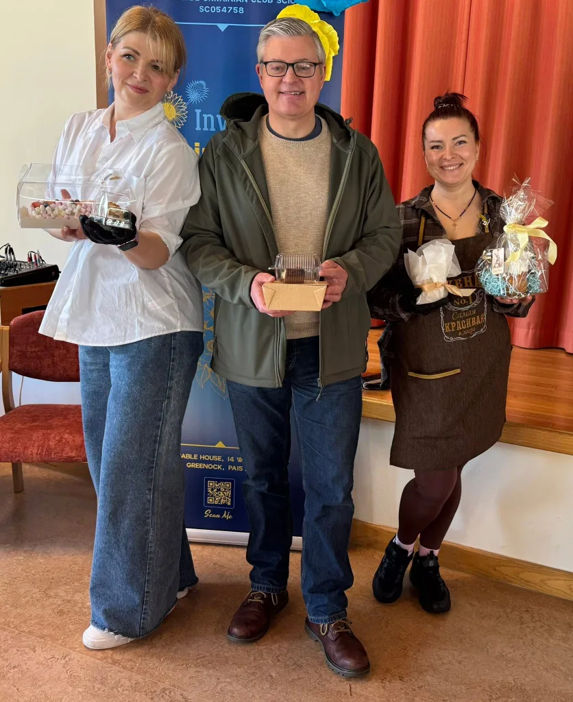 Three people stand together in front of a banner, holding boxes of Easter baking.