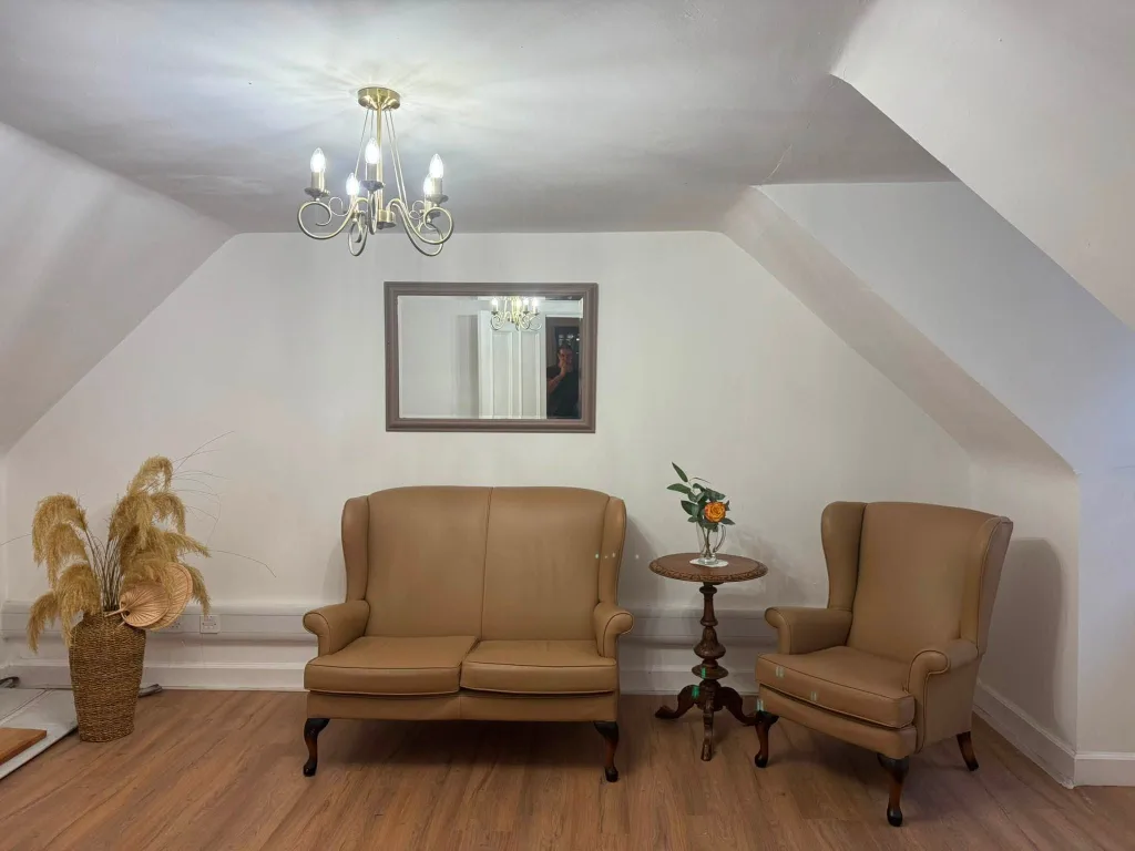 A sofa and a wing chair under the eaves, with a mirror above and flowers on a small table between them.