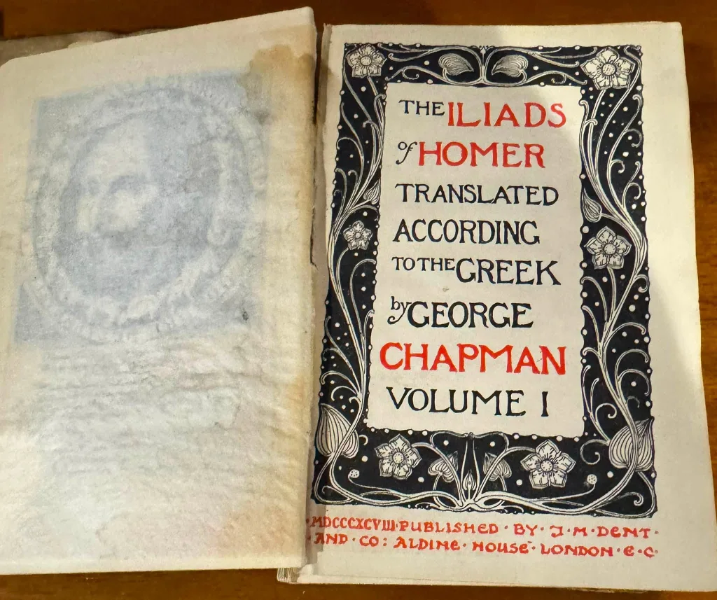 A book open at its title page: "The Iliads of Homer, translated according to the Greek by George Chapman, Volume I", published by J. M. Dent, Aldine House, London.