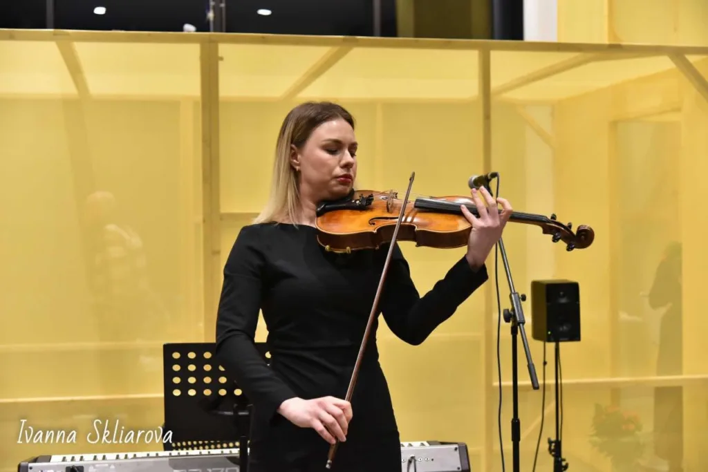 A violinist in black plays on a lit stage at a Club concert, a music stand and microphone beside her.