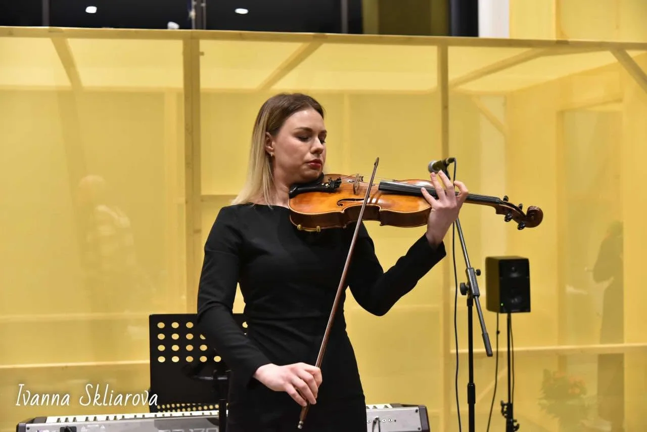 A violinist in black plays on a lit stage at a Club concert, a music stand and microphone beside her.