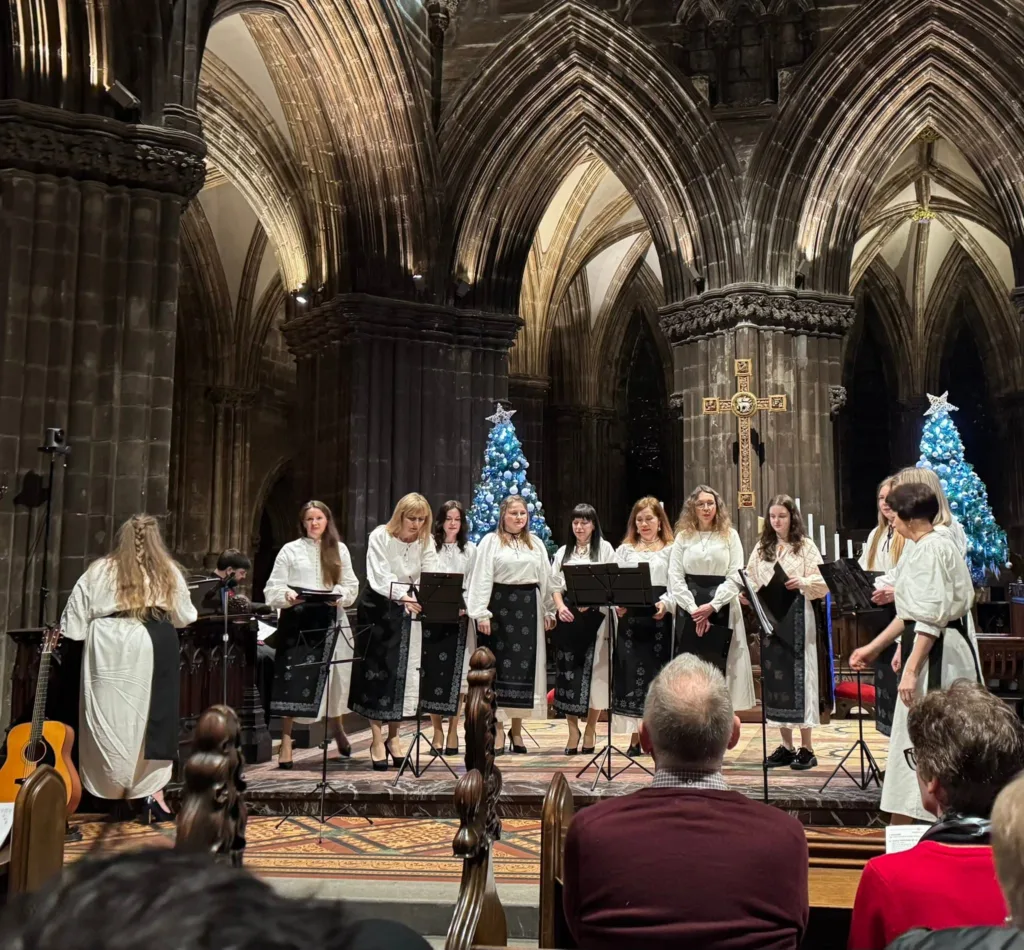 The Club's choir singing from music stands in a cathedral, in white embroidered blouses and dark patterned skirts, with lit Christmas trees behind them and an audience seated in front.