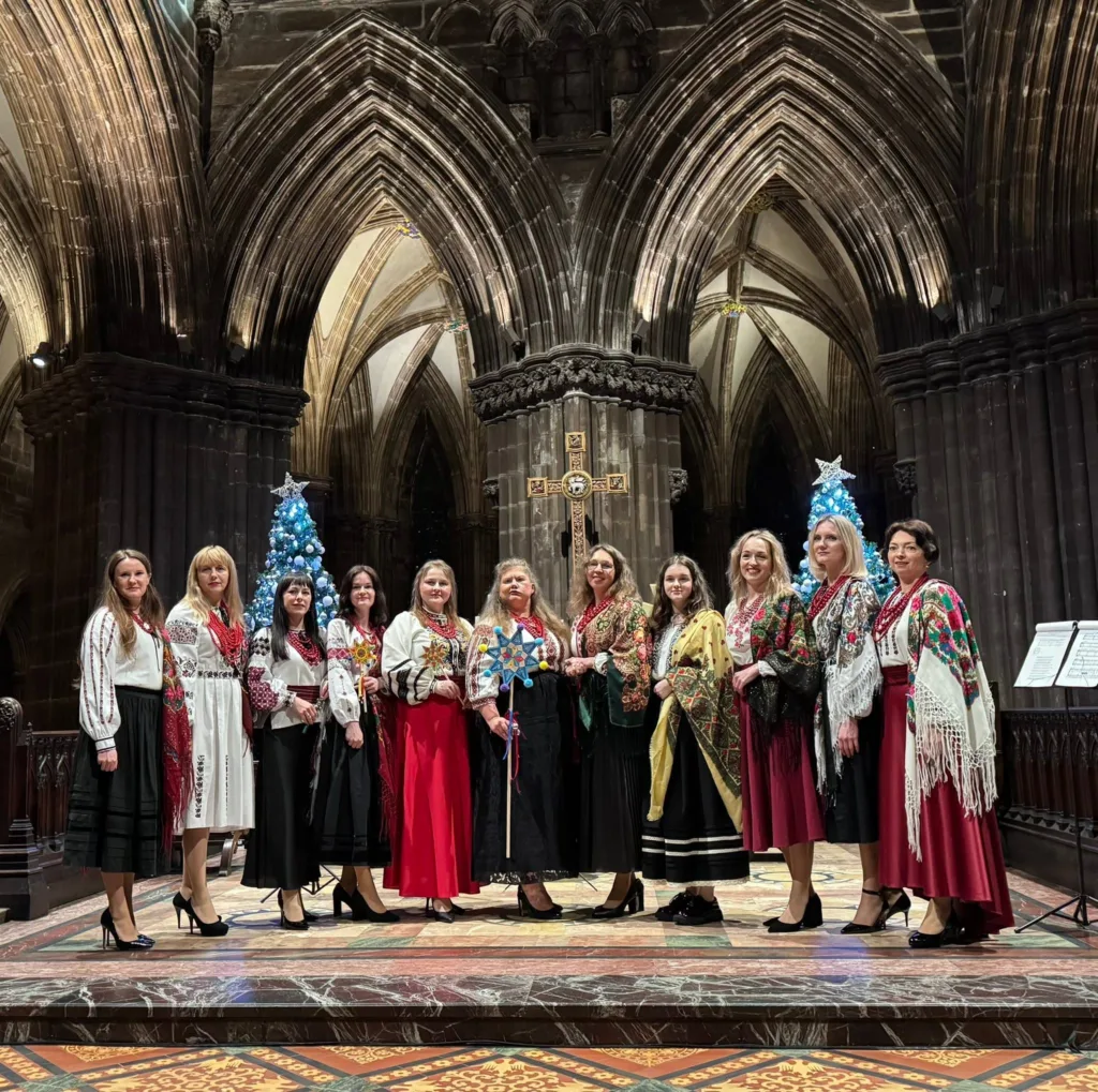 The Club's choir in embroidered vyshyvanky and patterned shawls, standing together before two lit Christmas trees in a cathedral. One of them holds a blue and yellow Christmas star on a pole.