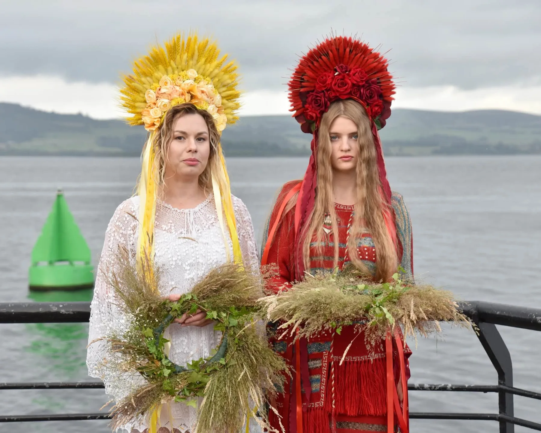 Two women in tall Ukrainian flower wreaths hold sheaves of wheat at the Gourock waterfront, with the Clyde behind them