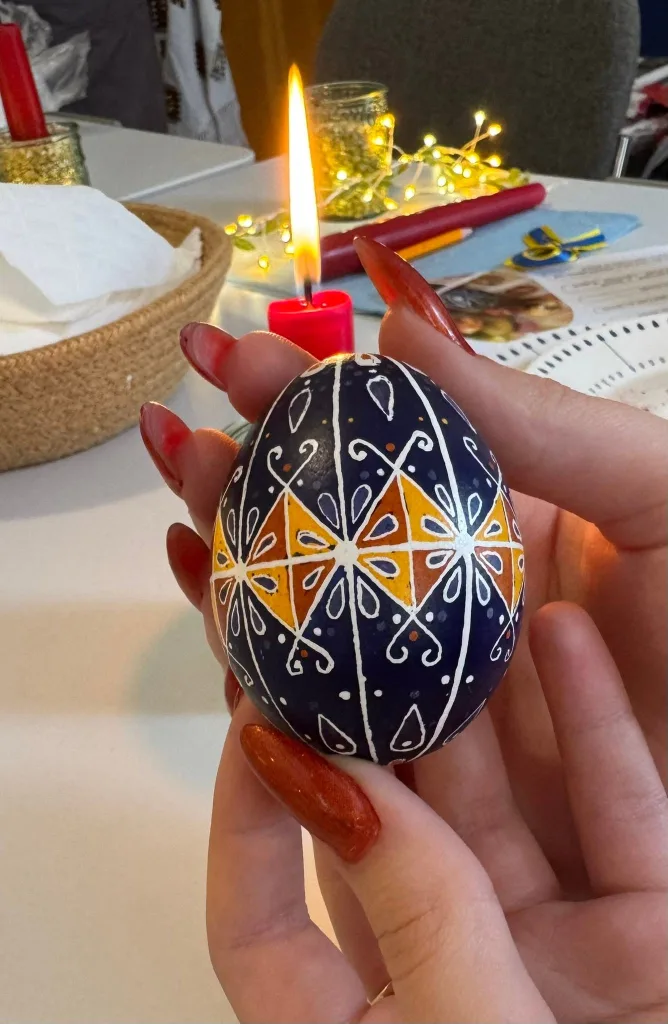 A hand holds a finished navy pysanka with a white and gold star pattern, a candle burning behind.