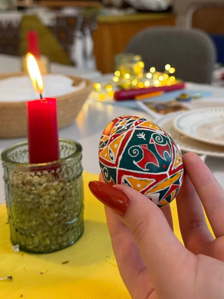 A hand holds a red, yellow and green pysanka beside a candle on a yellow cloth.