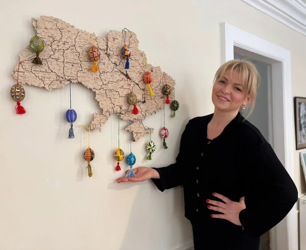 A woman stands beside a wooden map of Ukraine hung with pysanky, presenting it to the camera.