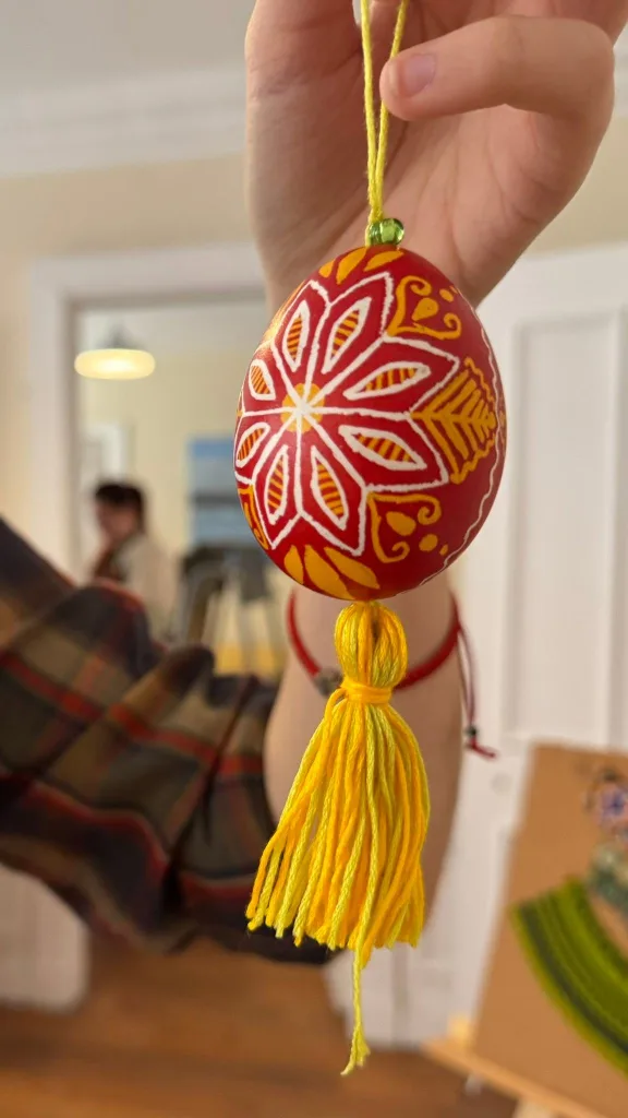 A red and white pysanka hanging from a yellow tassel, held up in a room.