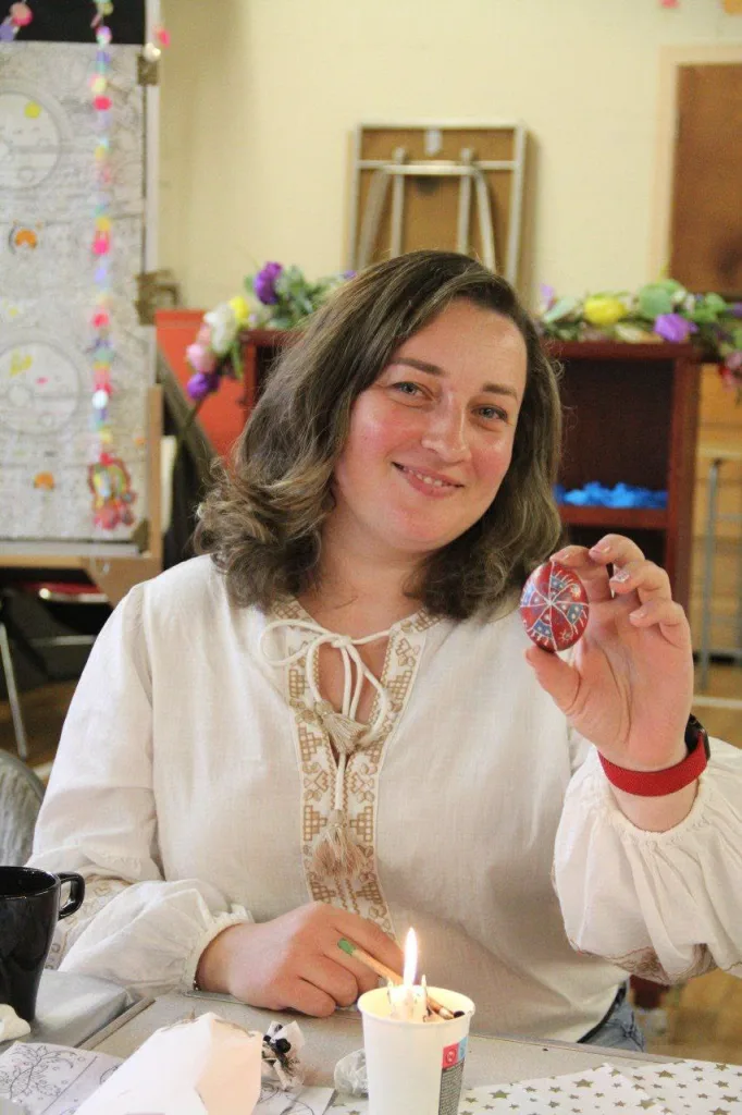 A woman in an embroidered shirt holds up a finished pysanka beside a candle.