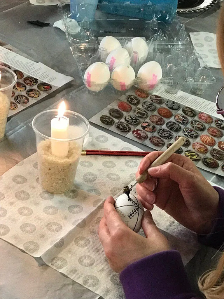 Hands drawing wax onto an egg with a kistka over a candle flame, trays of eggs behind.