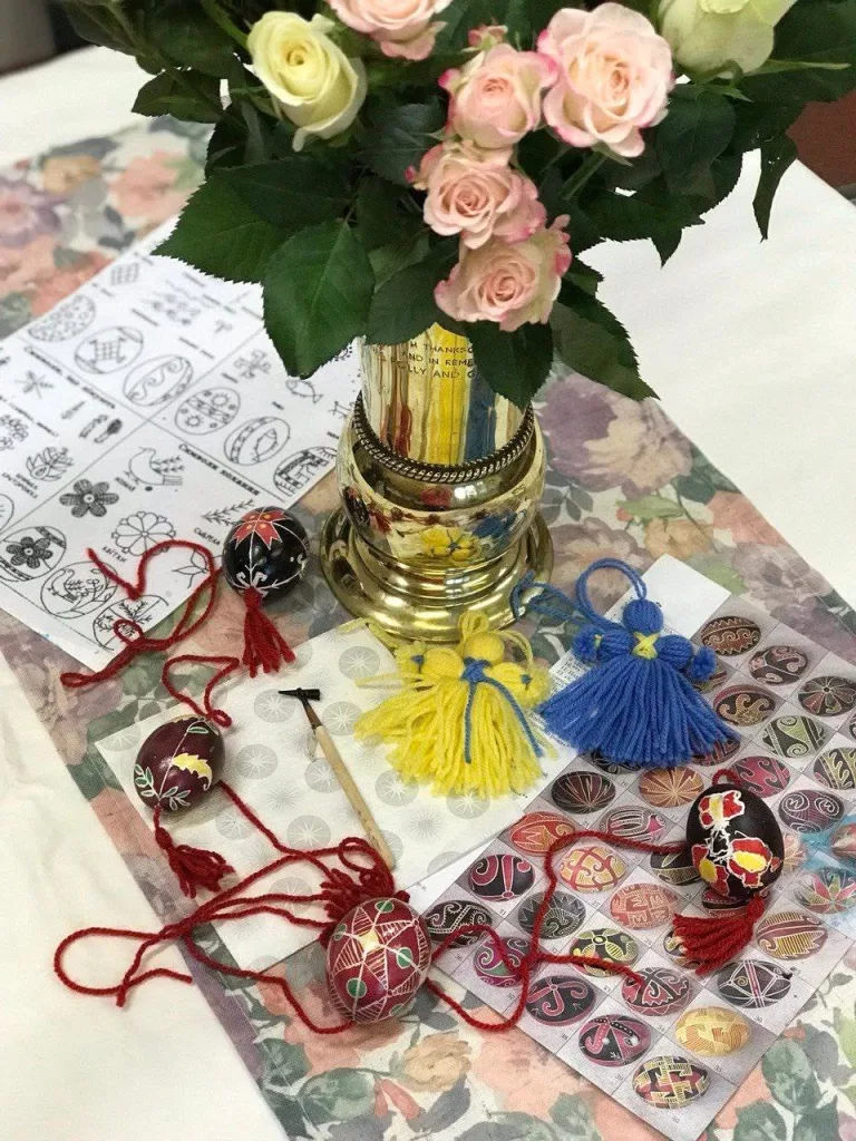 A vase of roses tied with blue and yellow ribbon, beside finished pysanky on tassels.